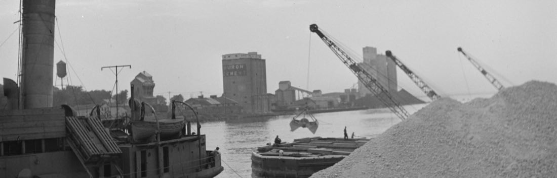 Bauxite being unloaded at the Port of Oswego in 1943: a freighter under steam at the wharf, three derrick cranes working a barge, and grain elevators on the skyline