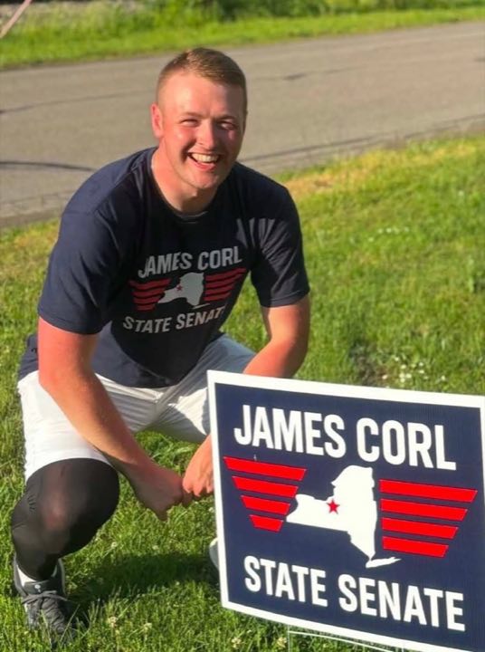 An Oswego County volunteer kneeling beside a James Corl for State Senate yard sign on a summer afternoon